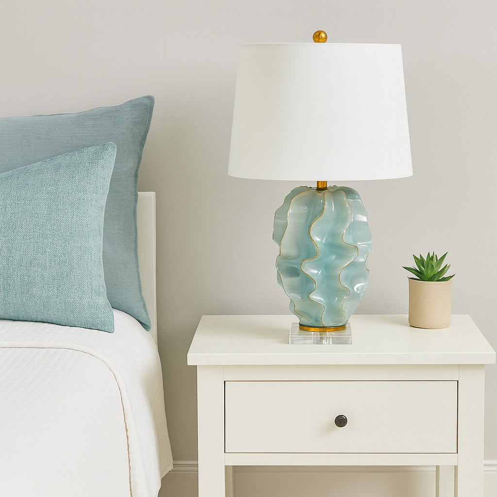 Table lamp with a decorative blue-green base on a nightstand next to a bed with light blue pillows.