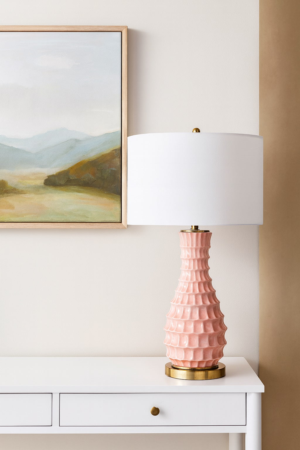 Pink textured table lamp with white shade on a white surface, with a framed landscape painting in the background.